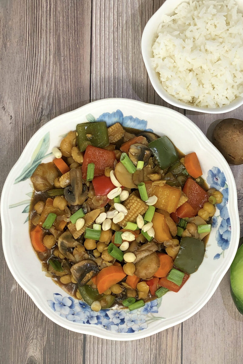chickpea vegetable kung pao in a bowl served with rice, half green pepper and mushroom in background.
