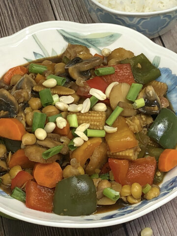 chickpea vegetable kung pao in a bowl served with rice, half green pepper and mushroom in background.